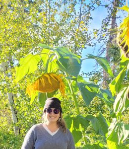 Image shows woman in a toque and sunglasses standing under a giant sunflower.