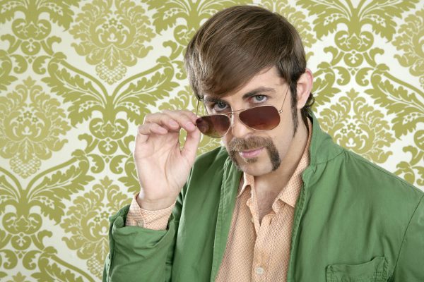 Image shows man with wearing 70s inspired clothing and facial hair standing in front of a retro wallpaper background