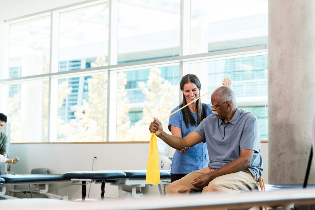 The smiling mid adult female activity assistant helps a senior man use the resistance band properly to strengthen his arm.