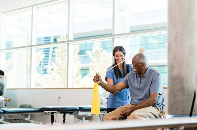 The smiling mid adult female activity assistant helps a senior man use the resistance band properly to strengthen his arm.