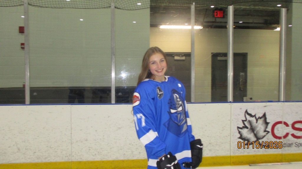 Image shows a young woman with long blonde hair wearing a hockey jersey on the ice.