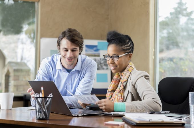 Diverse male and female business associates review documents together. They are looking at something on a laptop.