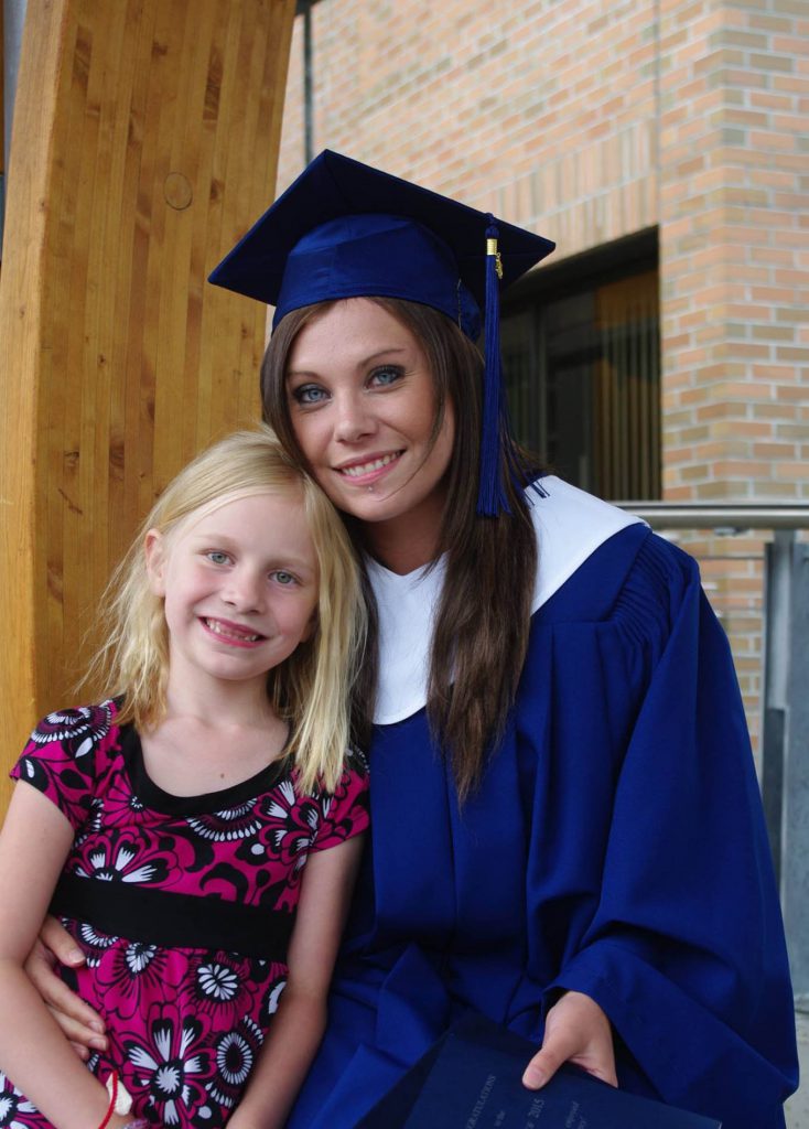 Image of dark-haired woman in graduation cap and gown with young blonde child on her lap.