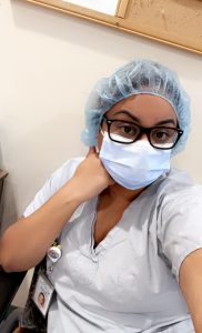 Photo of young woman in scrubs, mask, a hair covering. and dark glasses looks out at camera. 