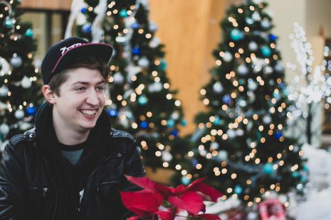 An image of a young man sitting in front of Christmas trees with a poinsettia in the forefront.