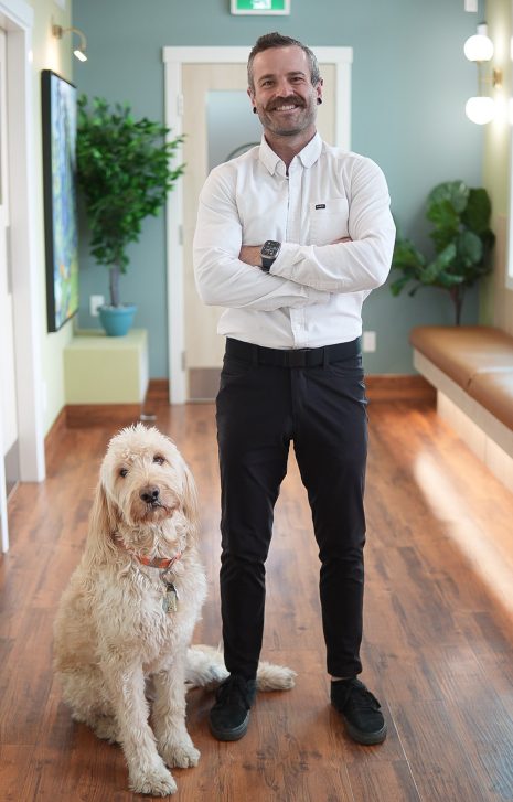 Image shows man smiling at the camera with a dog beside him