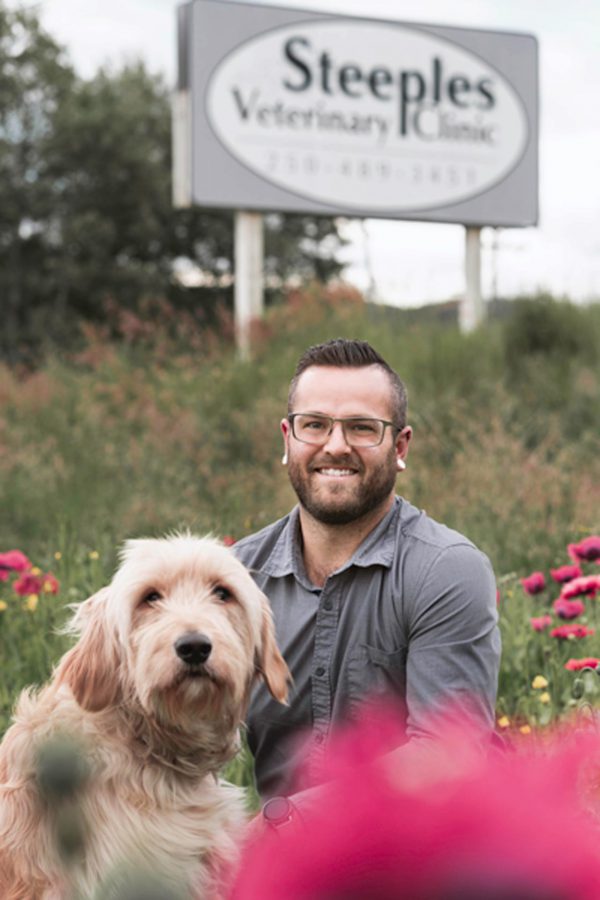 Image shows bearded man with glasses sitting in a field with a dog, with a Steeples Veterinary Clinic sign behind them.