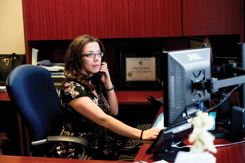 Image of woman in glasses sitting at a desk in front of a computer.