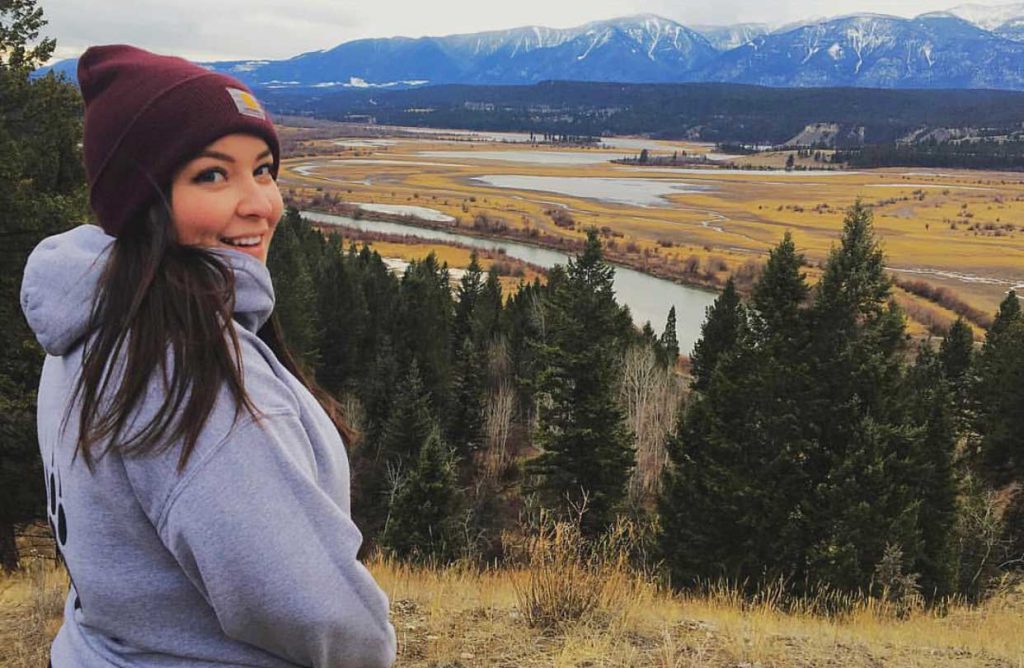 Image shows woman wearing a touque overlooking trees and water with mountains in the background.