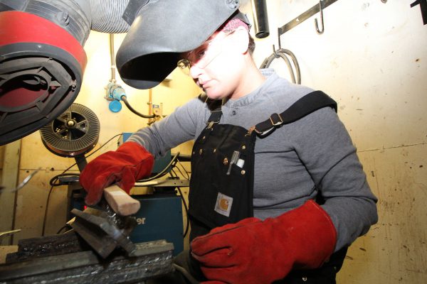 Image of woman in welding helmet, overalls, and welding gloves, cleaning a piece of metal with a wire brush.