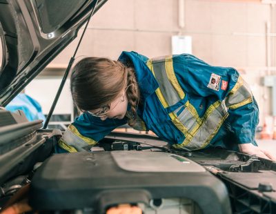 Image shows young woman working on the engine of a car.