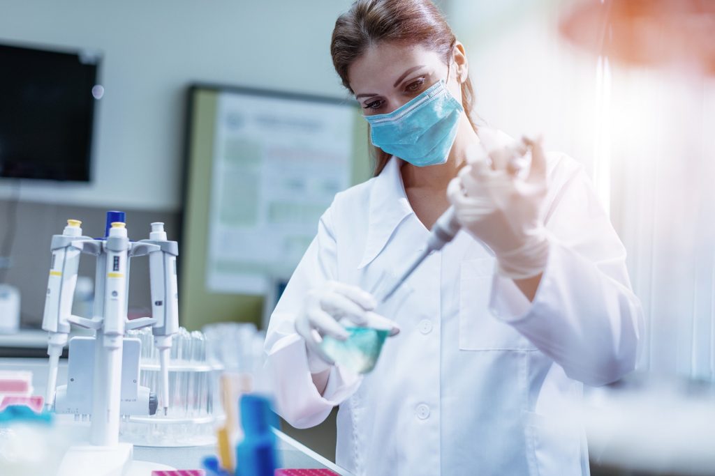 Woman in laboratory, examining new potions for the scientific research at university. Woman is wearing protective mask and protective gloves (Surgical gloves) while working with beakers, test tubes and other lab equipment.