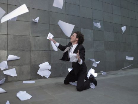 Image shows man in suit frantically trying to gather papers blowing in the wind.