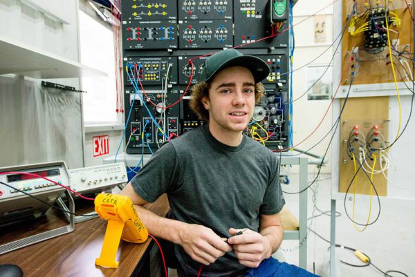 Image of young man sitting in front of a panel of wires and switches.