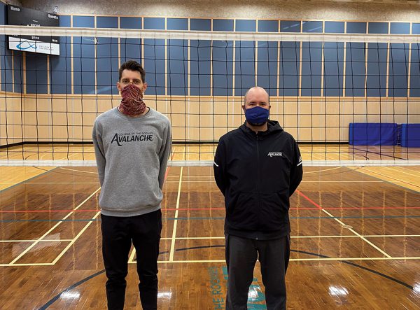 Image shows two men standing in front of a volleyball net in a gymnasium, both wearing masks.