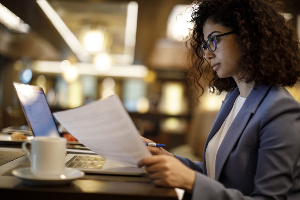 A woman sitting, looking at a document with a laptop computer in the background.