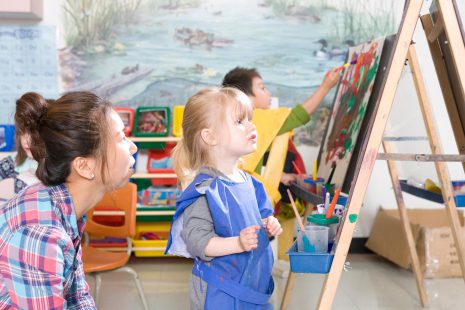 Image shows children painting at easels with a child care provider looking on and smiling.