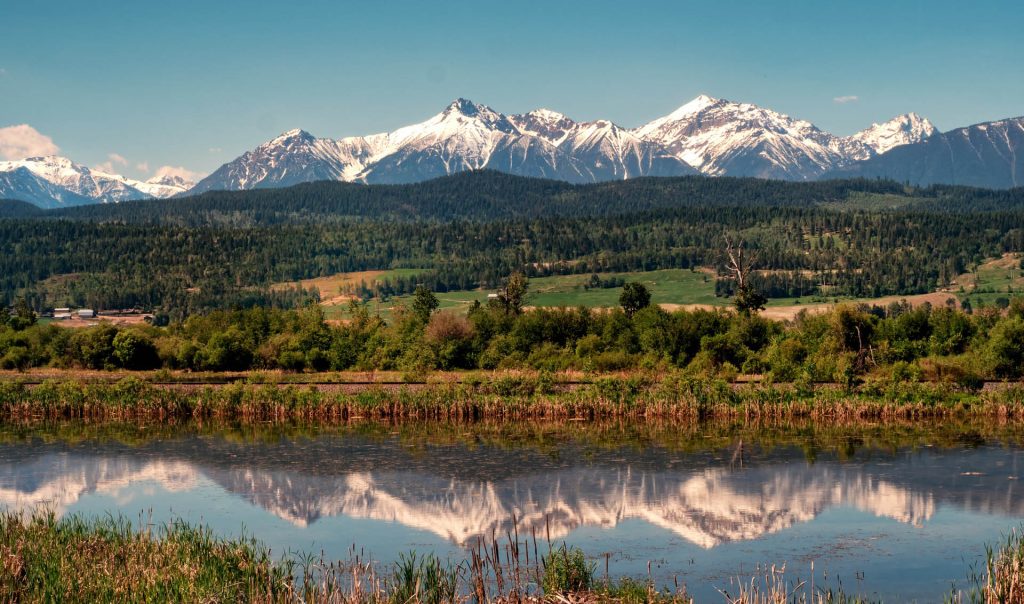 The snow capped mountains and wilderness contrast sharply with the tranquil Columbia river valley as it flows gently through the Rocky Mountain trench in western Canada