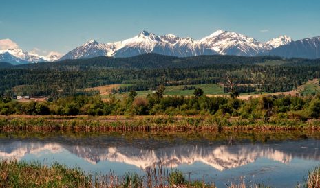 The snow capped mountains and wilderness contrast sharply with the tranquil Columbia river valley as it flows gently through the Rocky Mountain trench in western Canada