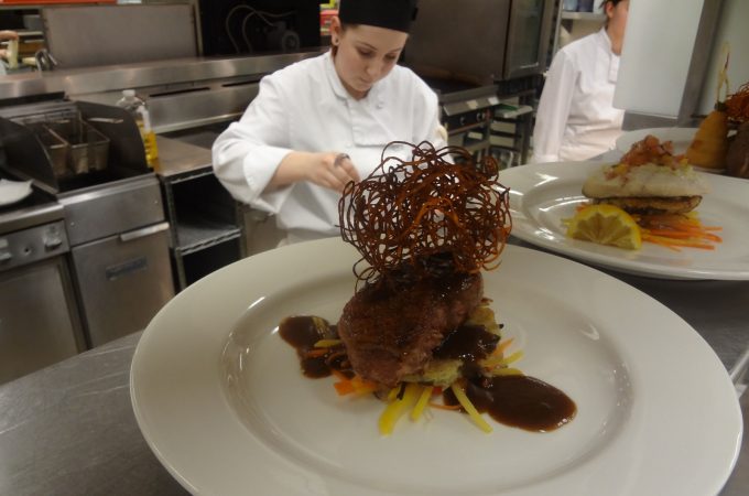 Cook training student in background with featured dish under heat lamp.