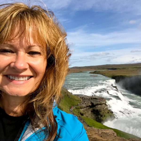 Image of smiling woman with windblown hair standing in front of Iclandic waterfall.