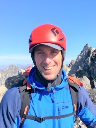 Image shows man outdoors with a backpack and helmet, smiling at the camera.