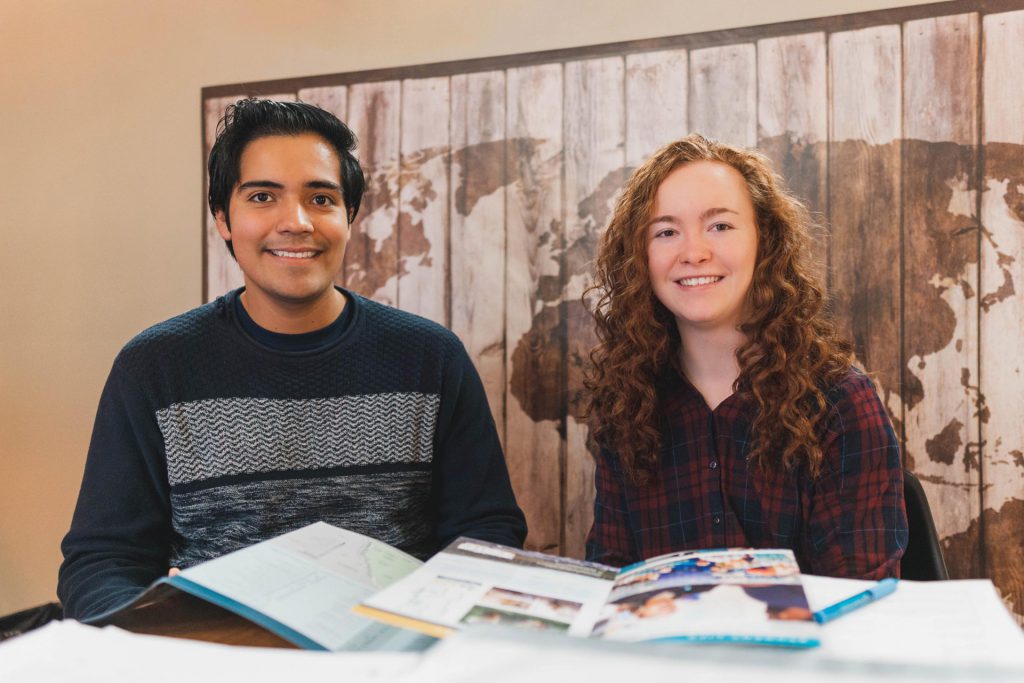 Image shows a young man and young woman sitting in front of a map of the world.