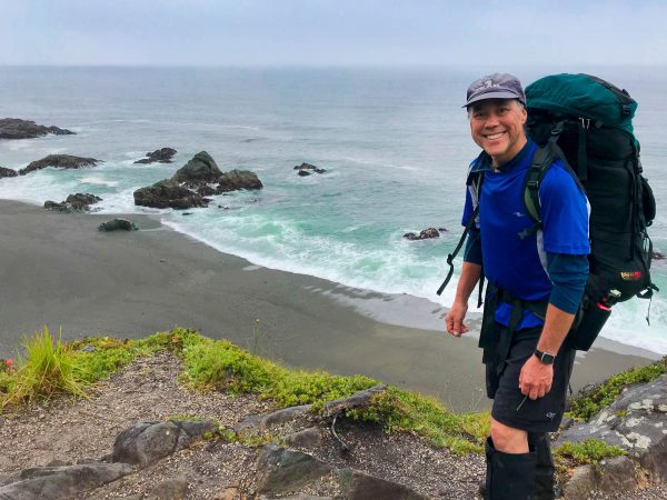 Image shows Dave Wan with large backpack on, hiking on the West Coast Trail.