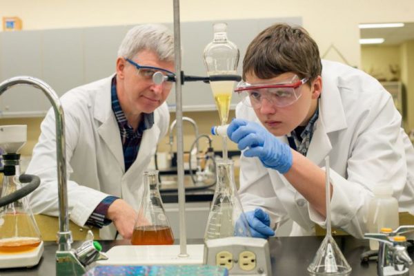 Image of man with gray hair, wearing a white lab coat and safety glasses, watching as a young man with similar attire manipulates liquid in beakers.