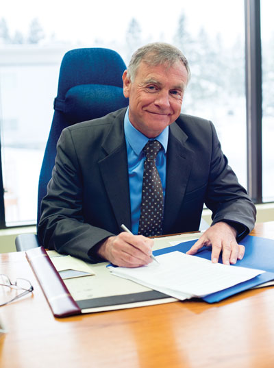 Image shows man sitting at desk with pen in hand and papers in front of him, smiling at the camera.