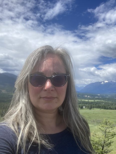 Image shows woman in sunglasses outdoor with mountains behind her.