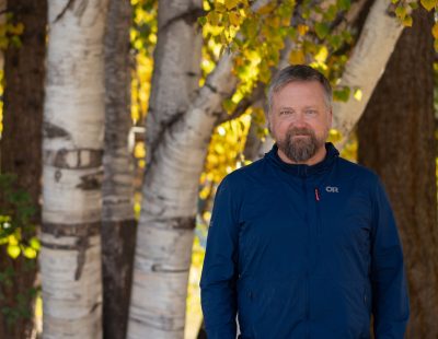 Image shows man standing in front of larch trees in the fall, looking at the camera.