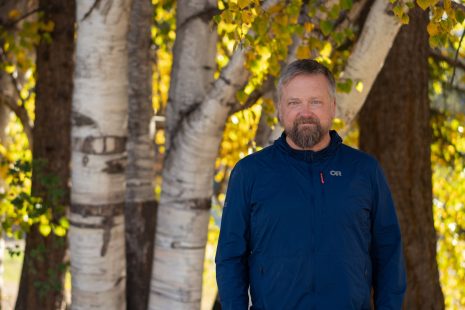 Image shows man standing in front of larch trees in the fall, looking at the camera.