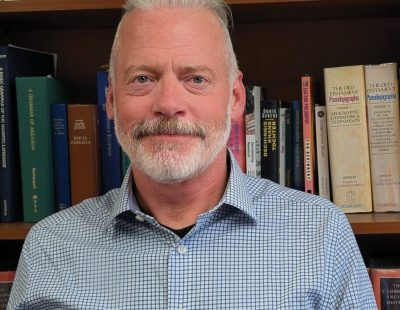 Image shows man with white hair and beard standing in front of a bookshelf.