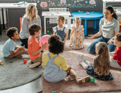 Image shows young students sitting in a circle with teachers.