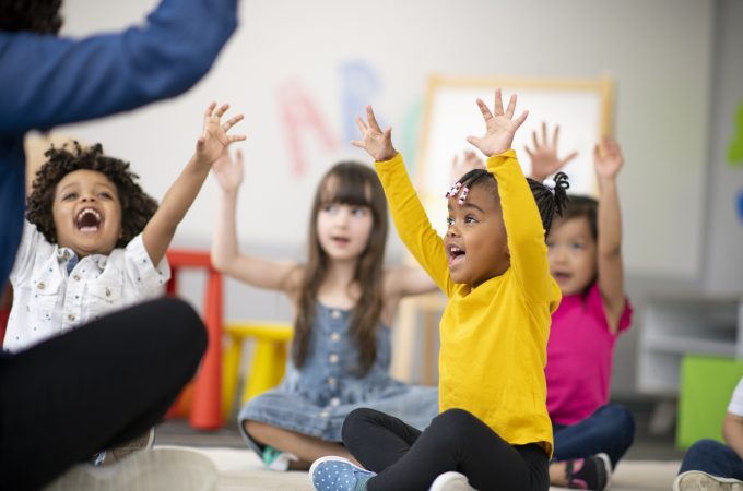 A multi-ethnic group of preschool students is sitting with their legs crossed on the floor in their classroom. The mixed-race female teacher is sitting on the floor facing the children. The happy kids are smiling and following the teacher's instructions. They have their arms raised in the air.
