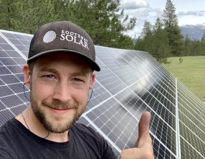 Image shows man in a Kootenay Solar baseball cap standing in front of a row of solar panels giving the thumbs up sign.