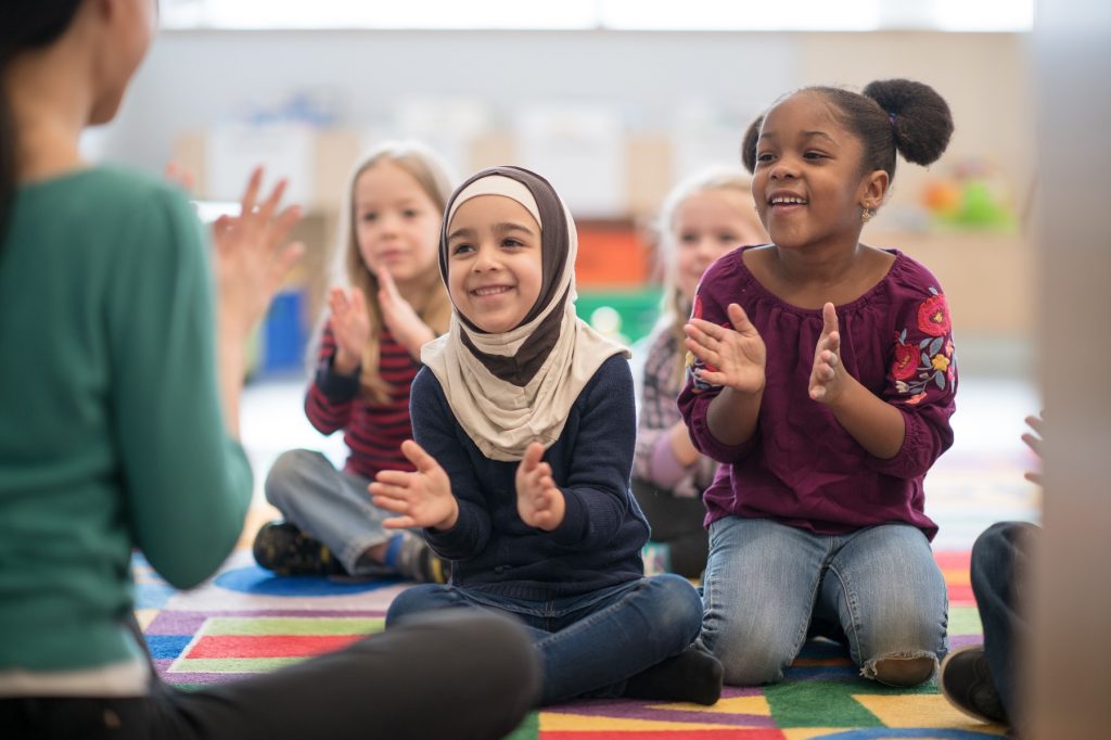 An adorable multi ethnic preschool children are singing and clapping their hands as their teacher is seated in front of them.
