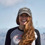 Image shows woman with long dark hair wearing a ball cap, with snow capped mountains behind her, smiling at the camera.