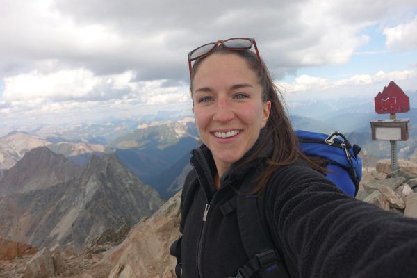 An image of a female standing on top of a mountain peak and smiling at the camera.