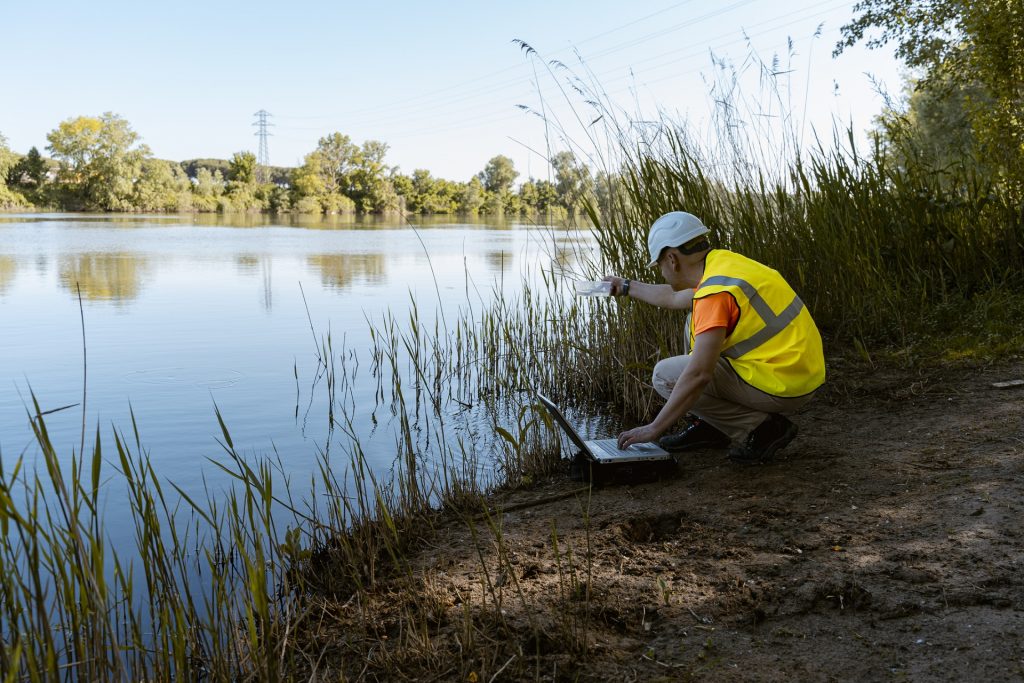 An environmental technician is crouched on the bank of a river, carefully collecting water samples. He is dressed in safety clothing, including a vest and helmet, indicating caution and professionalism in his work.