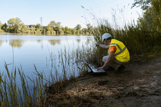 An environmental technician is crouched on the bank of a river, carefully collecting water samples. He is dressed in safety clothing, including a vest and helmet, indicating caution and professionalism in his work.