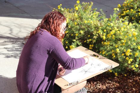 Image shows woman outside near a garden, sketching.