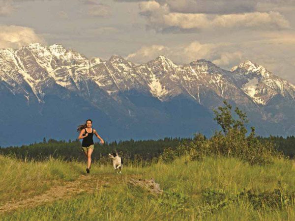 Image shows young woman running on a grassy path, with her dog, with snow-capped mountains in the background.