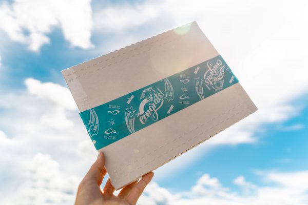 Image shows white box with teal packing tape with the words congrats written on it, being held up to a sunny sky.