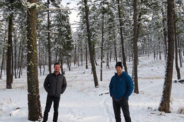 Image shows two men standing in snowy forest, simling at the camera.