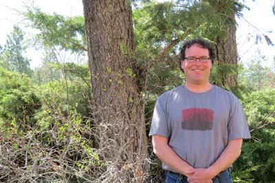 Image shows smiling man in glasses standing next to a tree and shrubs