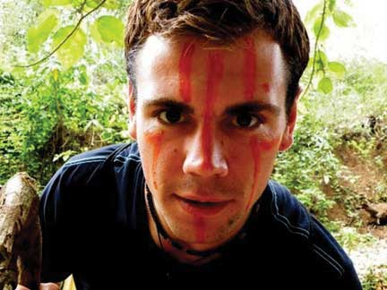 Image of Greg Snell in jungle-like setting with red paint on his face.