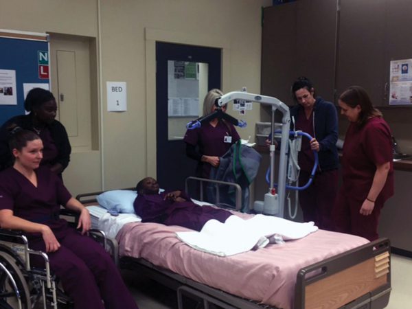 Five individuals in maroon scrubs, one in a wheelchair, surround individual lying on hospital bed.