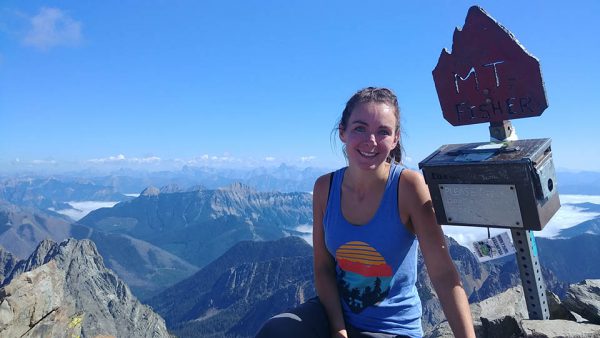 Image shows young woman smiling atop a mountain next to a sign reading Fisher Peak